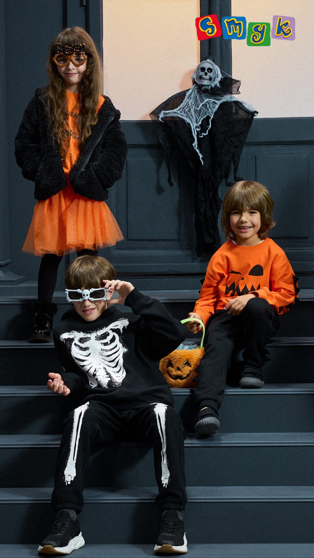 Czworo dzieci w halloweenowych stylizacjach z motywami dyni, szkieletu i kota, z pomarańczowymi torbami na słodycze. Grafika SMYK.