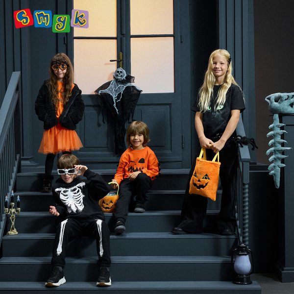 Czworo dzieci w halloweenowych stylizacjach z motywami dyni, szkieletu i kota, z pomarańczowymi torbami na słodycze. Grafika SMYK.