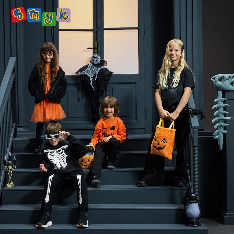 Czworo dzieci w halloweenowych stylizacjach z motywami dyni, szkieletu i kota, z pomarańczowymi torbami na słodycze. Grafika SMYK.