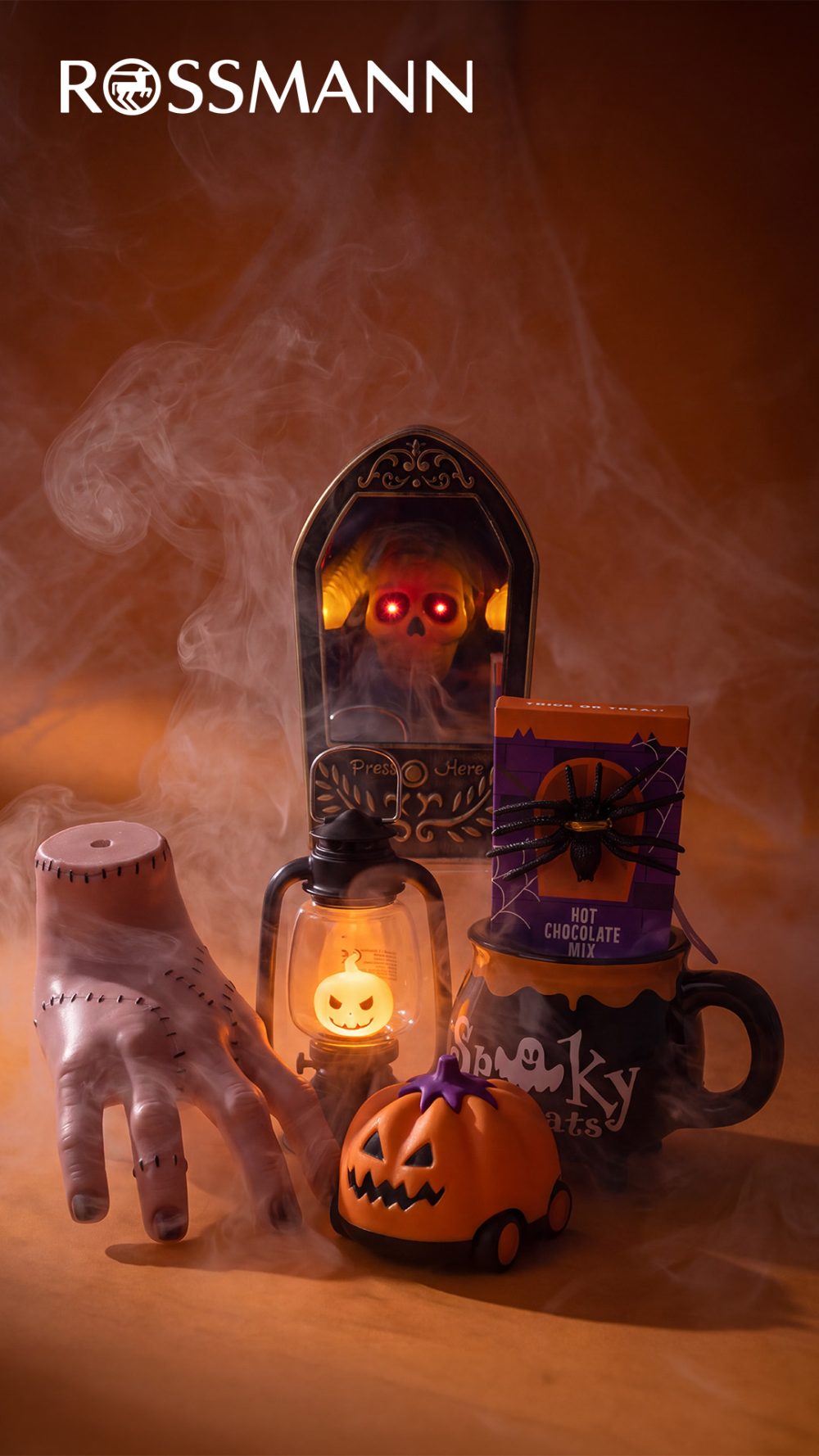 Dekoracje Halloween w Rossmannie: sztuczna ręka, dzwonek z czaszką, latarnia z dynią, pojazd w kształcie dyni oraz kubek z czekoladą do picia.